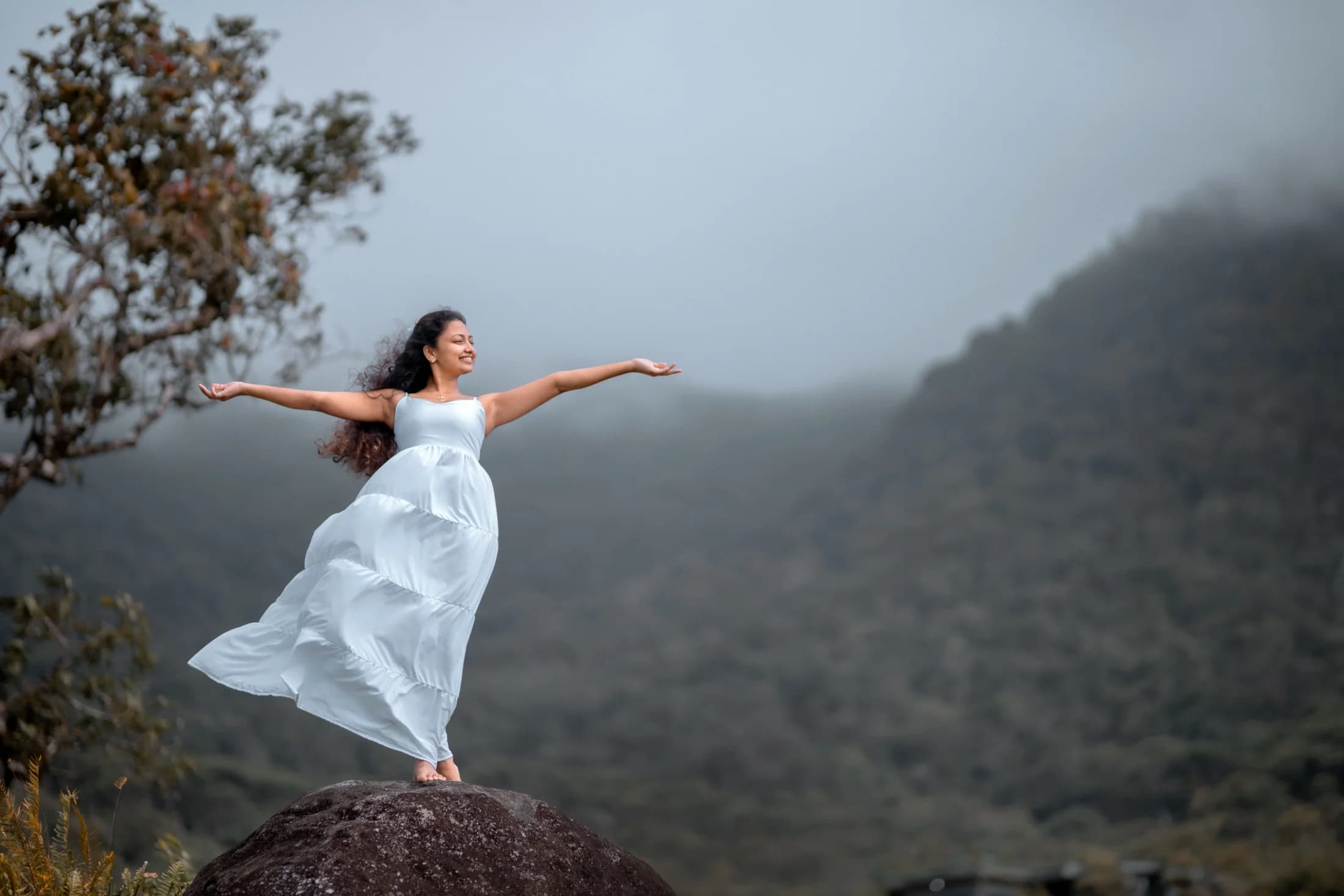 Woman in a white dress standing on a rock against misty mountains